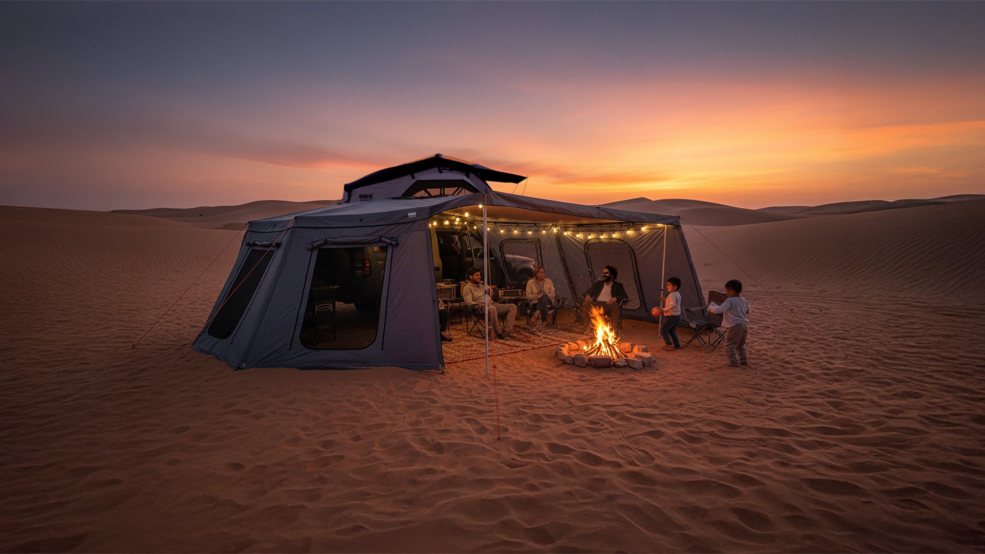 OBK 270 awning full setup in the desert at sunset, creating a majlis-style outdoor camping space with seating, warm string lights, a campfire, and family gathered underneath for a cozy evening atmosphere.